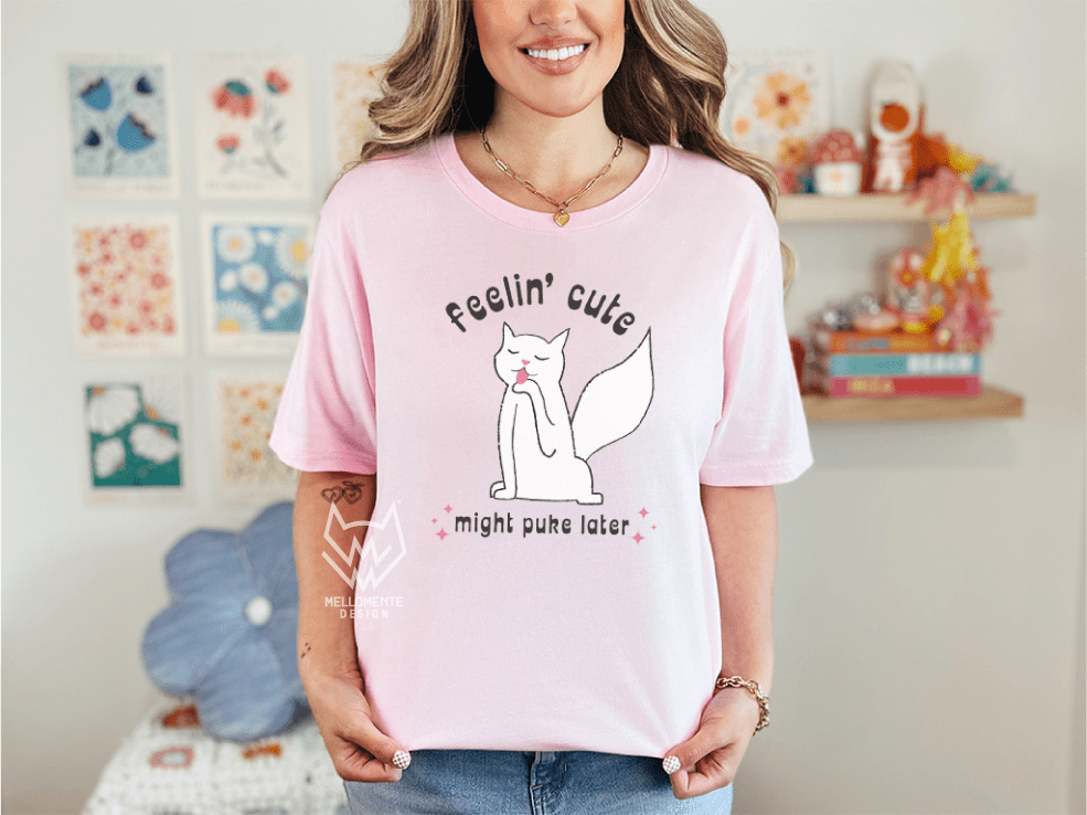 Person wearing a pink t-shirt with a white cat graphic and text, standing in a room with colorful decorations.